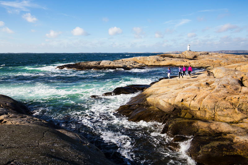 Group of people, with a lighthouse in the distance, hike on cliffs next to the sea - Photo cred Roger Borgelid