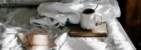 En närbild på en säng där det ligger böcker, en dagbok utspritt över lakanet. På en bok står en kopp kaffe